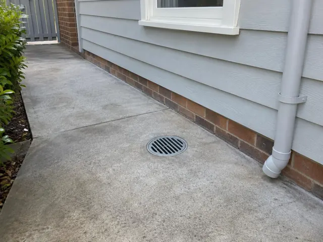 Outdoor concrete side path with a round drain grate beside a house wall, dry and clean in Gordon