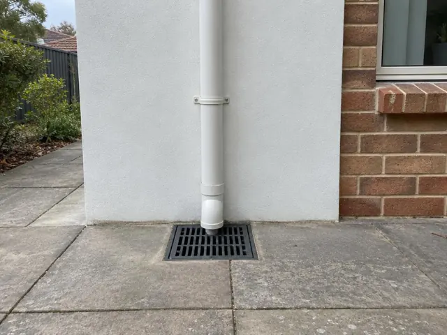 Stormwater grate beneath a downpipe beside brick and rendered house walls with dry paving in St Ives
